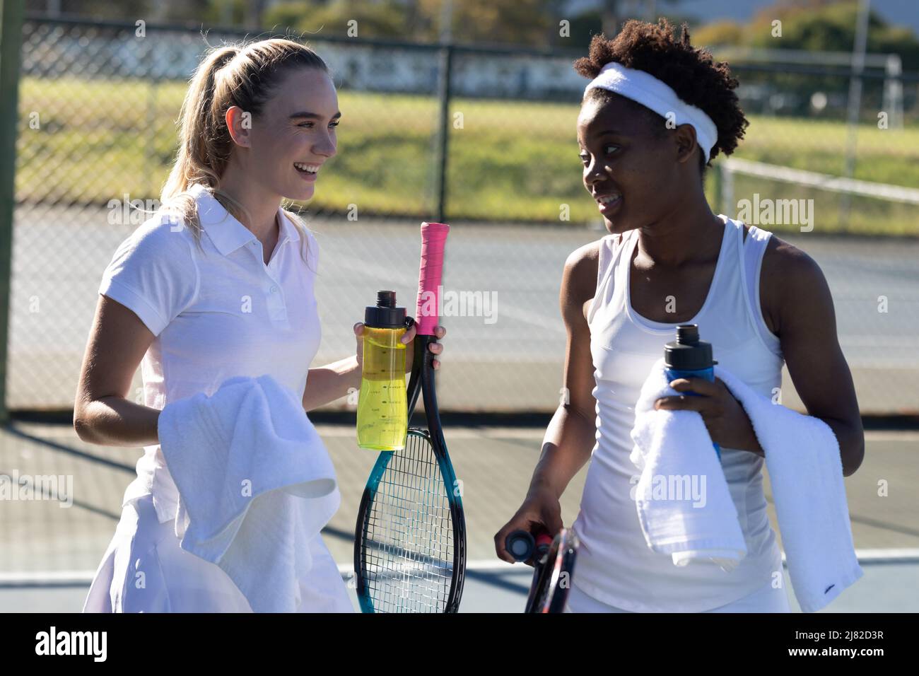 Des femmes souriantes de tennis biracial avec des bouteilles et des serviettes parlent pendant les pauses sur le court Banque D'Images