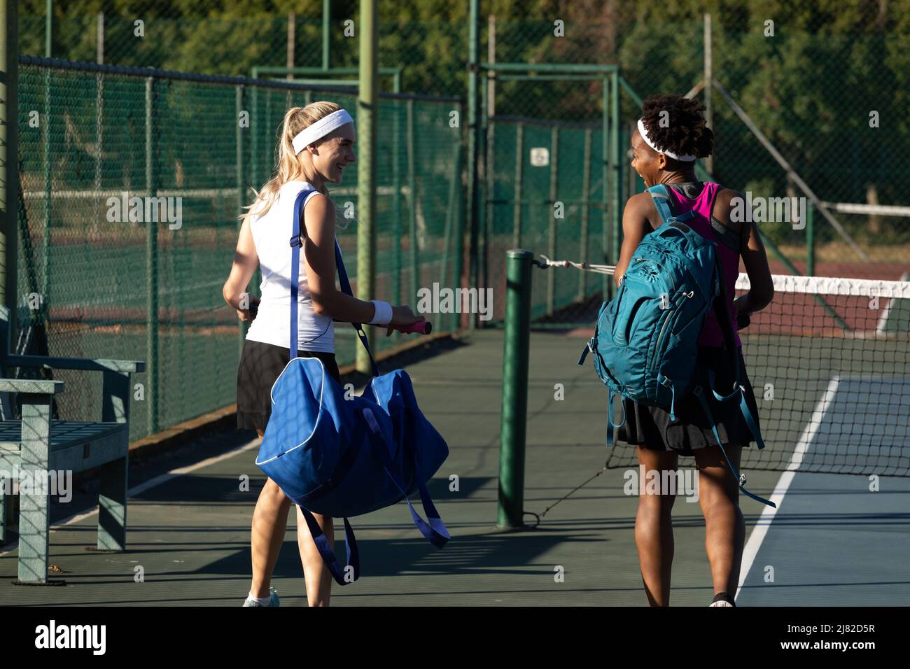 Souriante, jeune femme de tennis multiraciale marchant avec des sacs sur le terrain par beau temps Banque D'Images