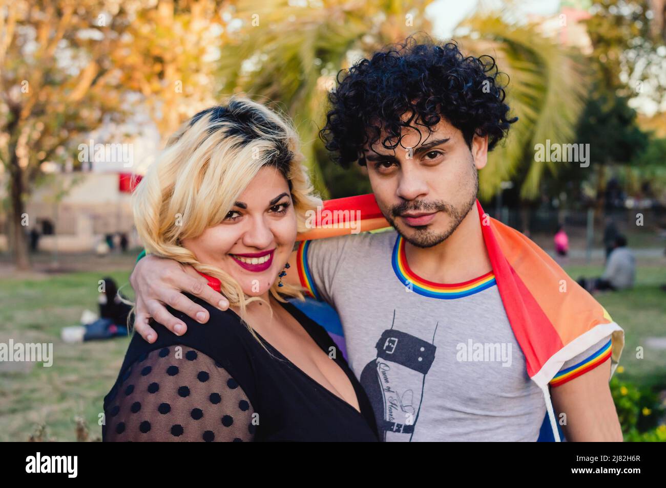 homosexuel homme caucasien, cheveux bouclés, avec le drapeau de fierté gay, hale sa femme ami à l'extérieur, regardant la caméra. Banque D'Images