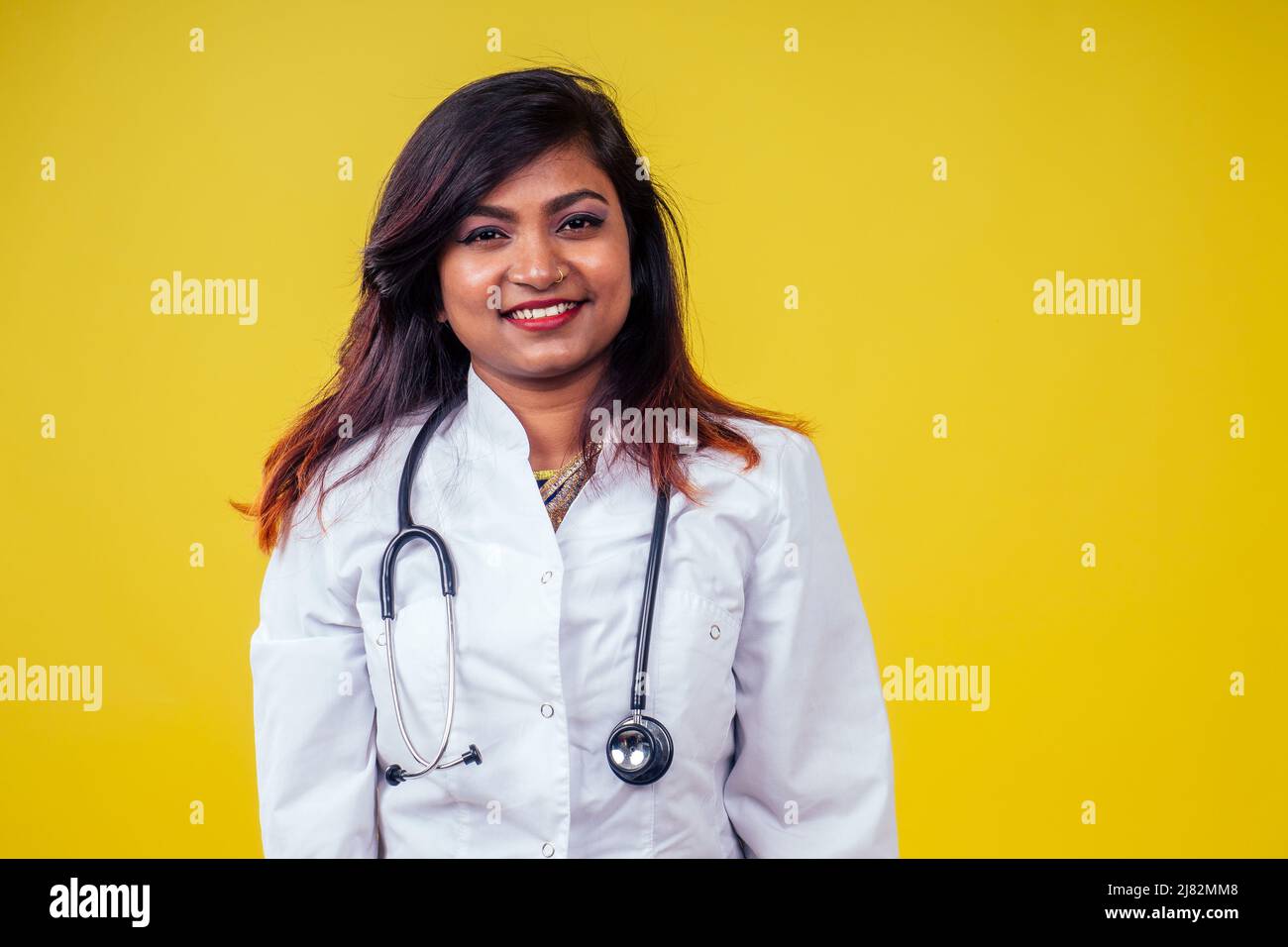 Femme indienne jeune et belle blonde femme gynécologue médecin utilisant le stéthoscope dans un manteau médical blanc sur un fond jaune dans le studio Banque D'Images