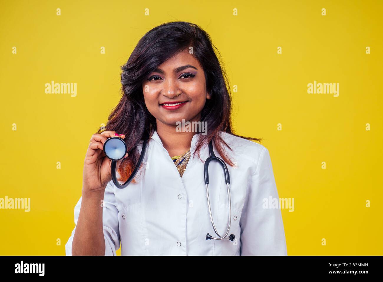 Femme indienne jeune et belle blonde femme gynécologue médecin utilisant le stéthoscope dans un manteau médical blanc sur un fond jaune dans le studio Banque D'Images
