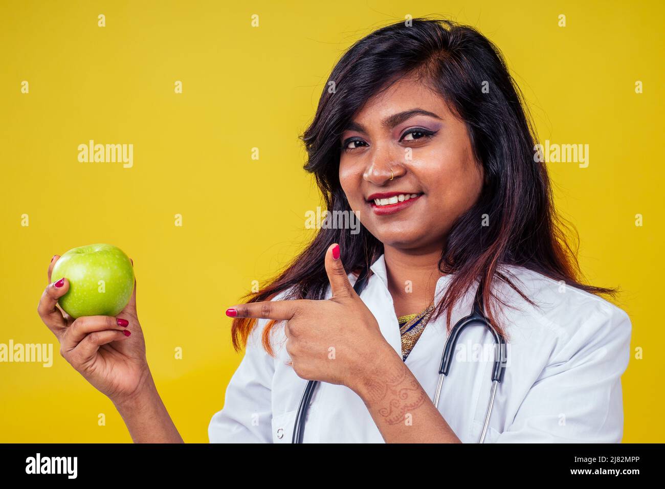Femme indienne jeune et belle blonde femme gynécologue médecin utilisant le stéthoscope tenant une pomme verte dans la main dans un manteau médical blanc sur un jaune Banque D'Images