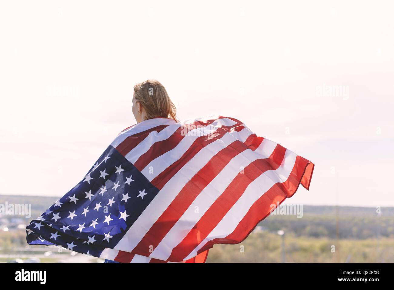 Jeune belle femme portant le drapeau des États-Unis. Le dos d'une jeune femme avec le drapeau national des États-Unis sur ses épaules, sur fond de montagnes cel Banque D'Images