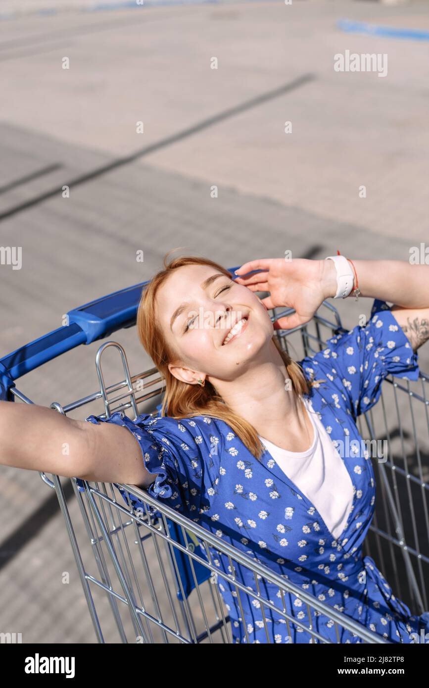 Une jeune femme gaie et élégante vêtue d'une robe bleue par temps ensoleillé se trouve dans un chariot de magasinage en face d'un supermarché. Mode cool sourire fille. Banque D'Images