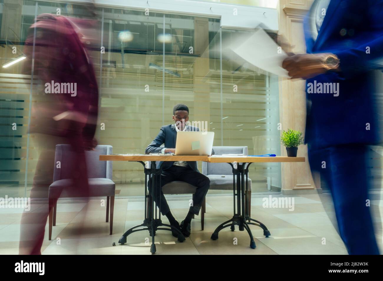 homme d'affaires américain d'afrique travaillant avec un ordinateur portable, personnes floues marchant à côté de lui dans un grand bureau avec de grandes fenêtres Banque D'Images