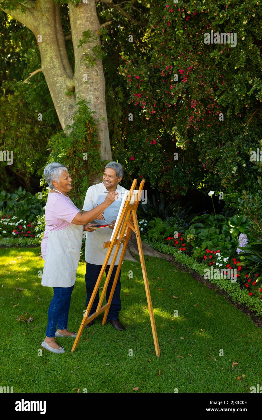 Homme biracial senior regardant la femme heureuse peindre sur toile tout en se tenant contre les plantes dans la cour Banque D'Images