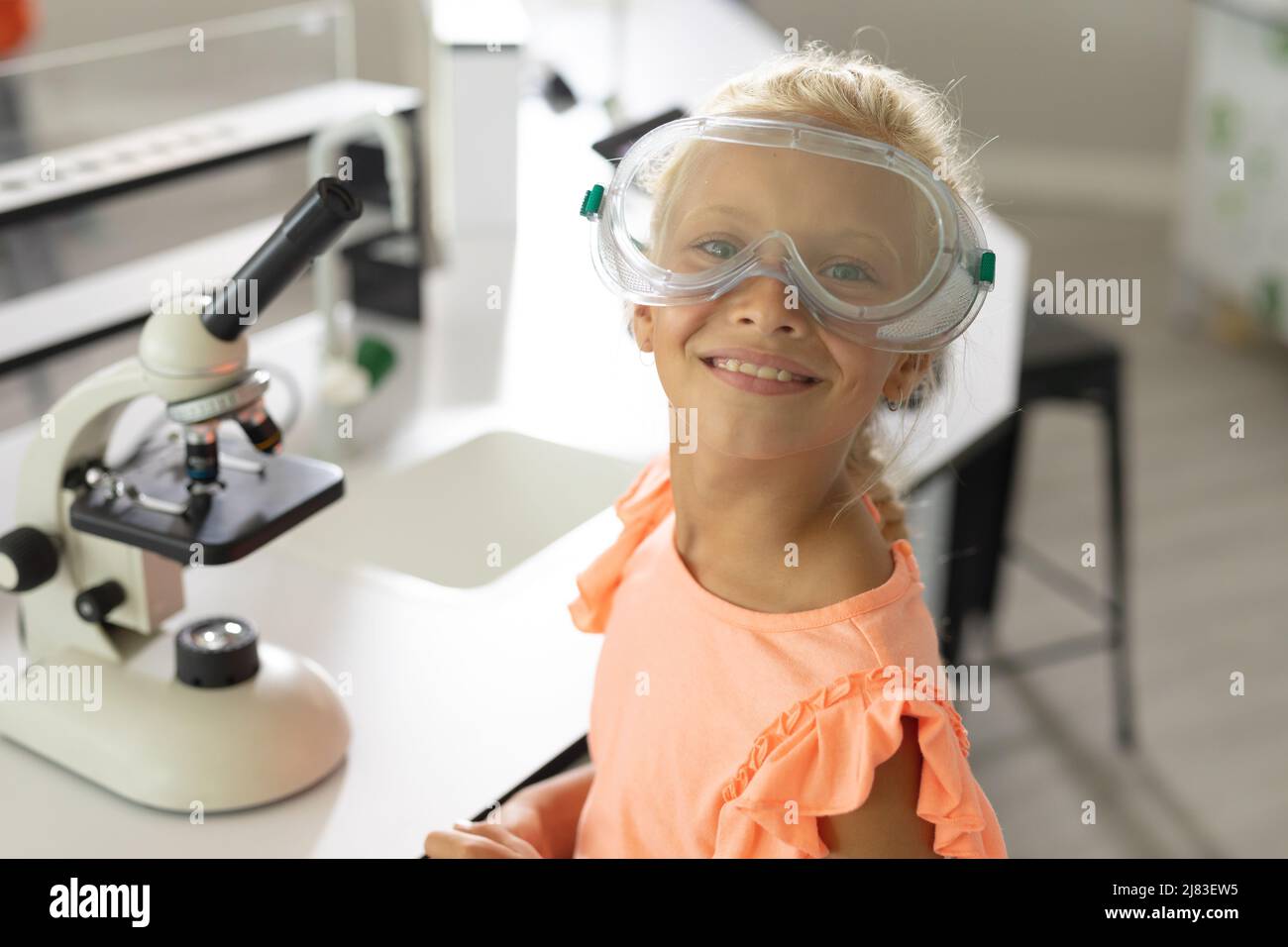 Portrait d'une jeune fille d'école élémentaire caucasienne souriante avec des lunettes de protection assises en laboratoire Banque D'Images