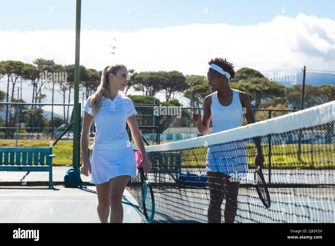 Souriant, de jeunes joueuses de tennis multiraciales parlent tout en marchant sur le court par beau temps Banque D'Images