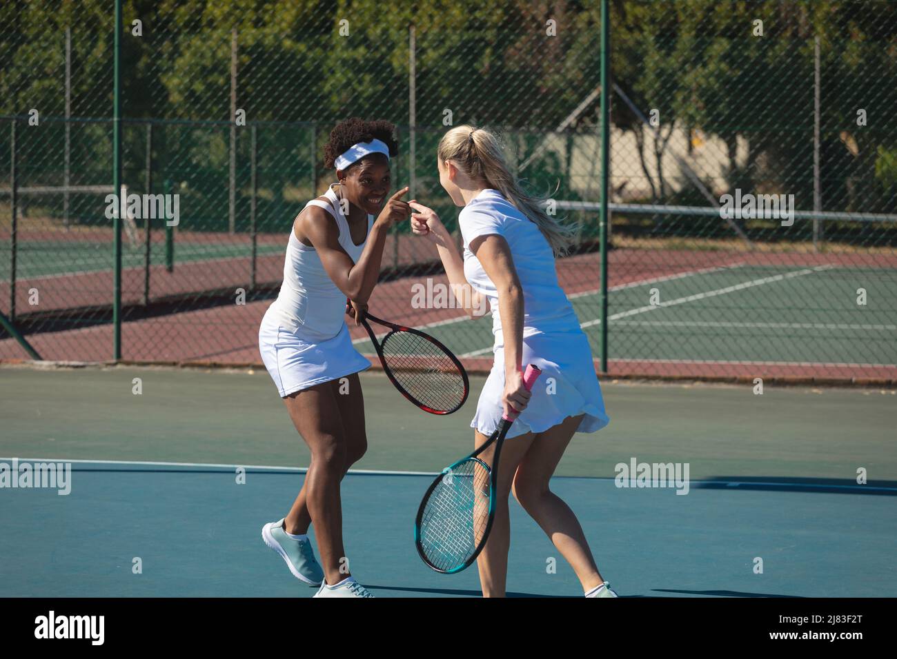 Des joueuses de tennis biraciales gaies se pointant les unes vers les autres tout en jouant sur le terrain par beau temps Banque D'Images