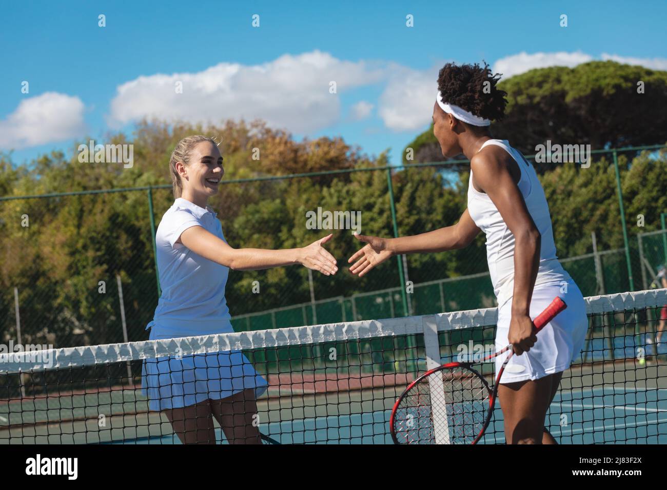 Des joueuses de tennis multiraciales heurtant les mains sur le filet sur le court par beau temps Banque D'Images
