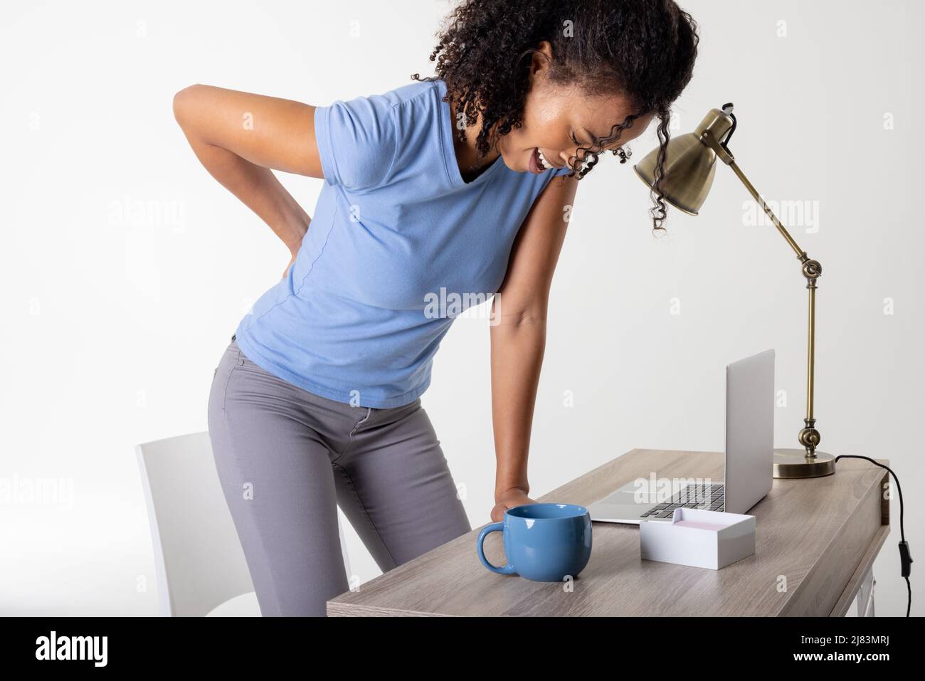Afro-américaine femme adulte de taille moyenne avec mal de dos debout par ordinateur portable, tasse et lampe à la table, espace de copie Banque D'Images