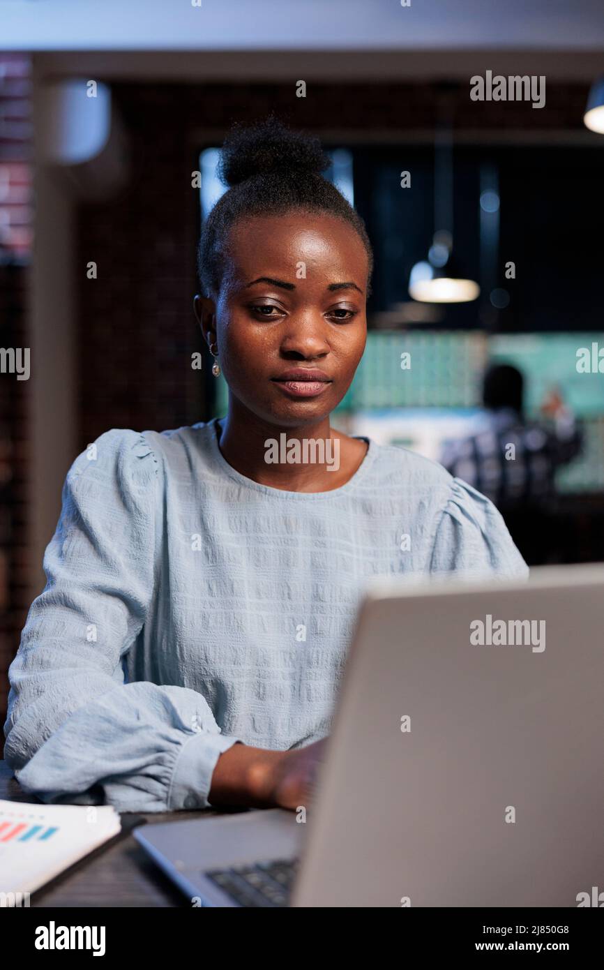 Portrait d'une femme afro-américaine confiante assise à un bureau tout en utilisant un ordinateur portable pour effectuer une analyse de marché en temps réel. Trader de stock de Forex dans l'espace de travail d'agence examinant les statistiques d'investissement. Banque D'Images