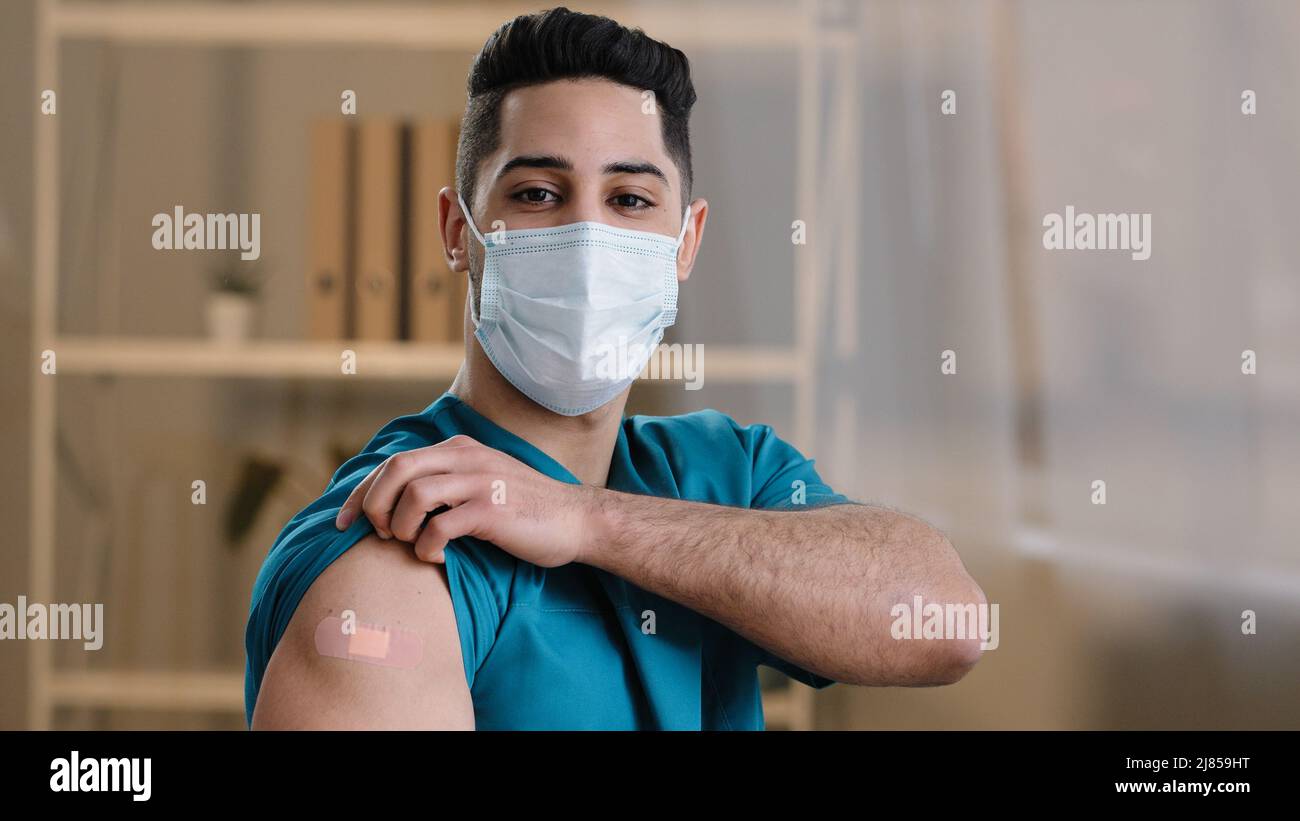 Jeune homme arabe praticien médecin infirmière debout à l'intérieur dans le masque chirurgical protecteur de visage démontrer le bandage adhésif sur l'épaule après le vaccin Banque D'Images