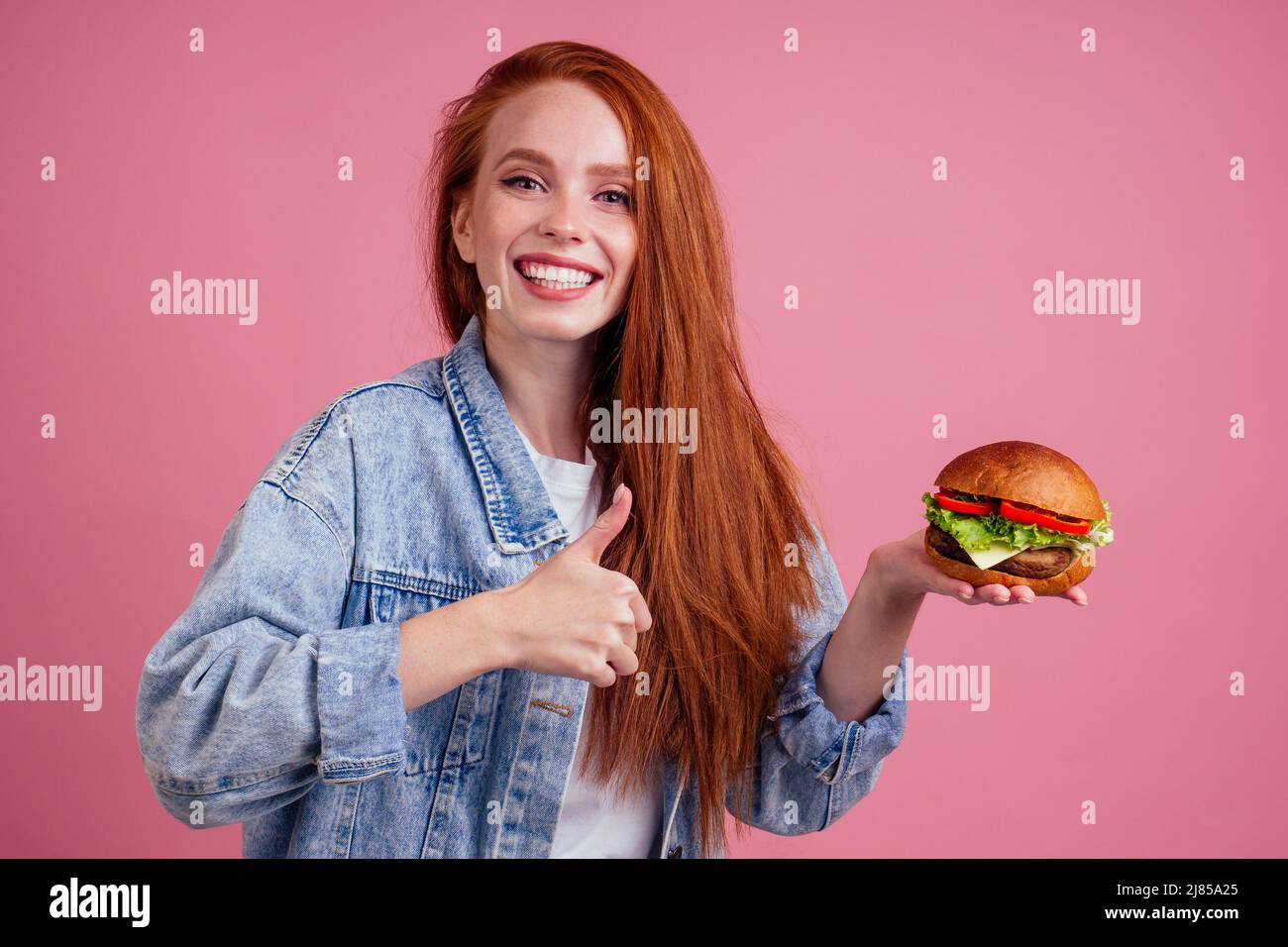 belle femme à tête rouge au gingembre, tenant un gros cheeseburger avec du fromage, des côtelettes de bœuf et de la laitue tomate sur fond rose studio Banque D'Images