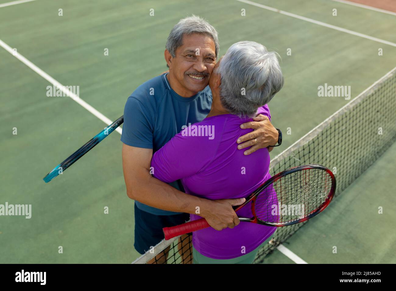 Portrait en grand angle de l'homme aîné biracial embrassant la femme tout en se tenant près du filet de tennis dans le court Banque D'Images