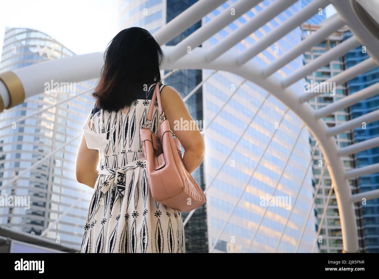 La vue arrière d'une jeune femme d'affaires marchant sur un pont supérieur d'intersection dans le centre-ville de Bangkok avec des immeubles de bureaux en hauteur en arrière-plan. Banque D'Images