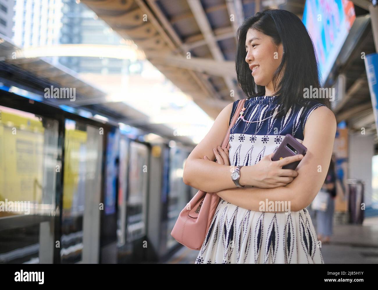 Une femme d'affaires asiatique vêtue de blanc et de bleu est debout avec ses bras croisés, attendant que le train aérien à la gare se rendent au travail dans la mornine Banque D'Images