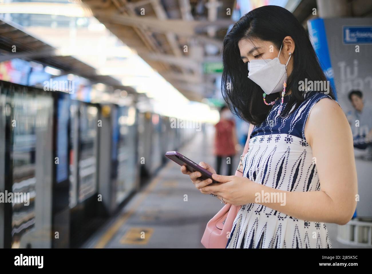 Une femme d'affaires attirante avec un masque de visage utilise son téléphone portable tout en attendant le train à une gare pour aller au travail le matin. Masque facial pour Banque D'Images