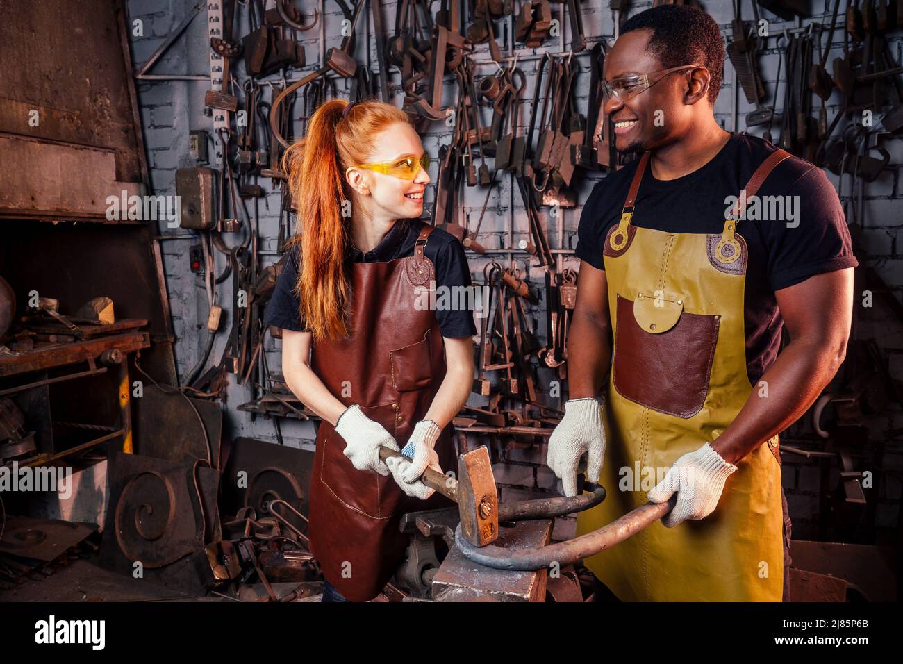 couple dans l'amour redhaired gingembre jeune femme européenne et afro-américain homme portant le tablier de cuir travail forgeron atelier.petite famille Banque D'Images