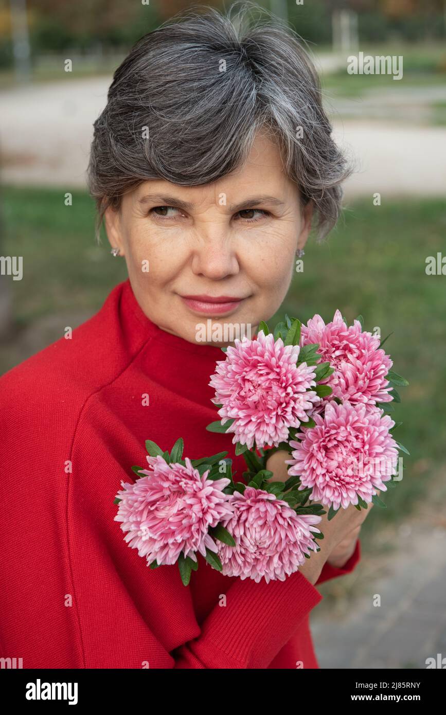 Portrait semi-profil d'une jolie femme à cheveux gris dans un chemisier rouge avec un bouquet de chrysanthèmes violets, regardant d'un côté. Taille haute. Banque D'Images