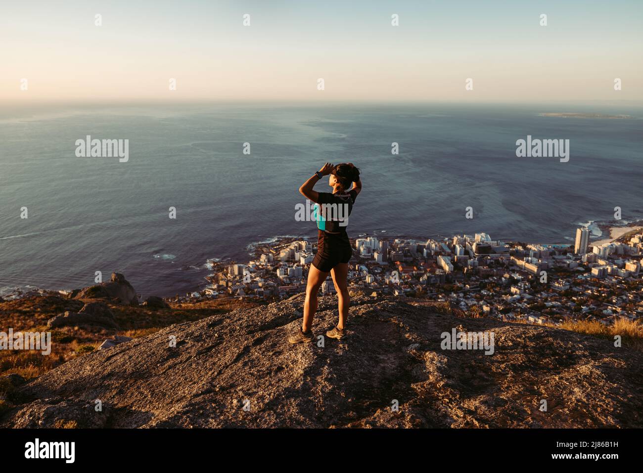 Femme en vêtements de sport debout sur la falaise et regardant la vue au coucher du soleil. Femme de randonnée se détendant sur le sommet de la montagne en regardant l'océan. Banque D'Images