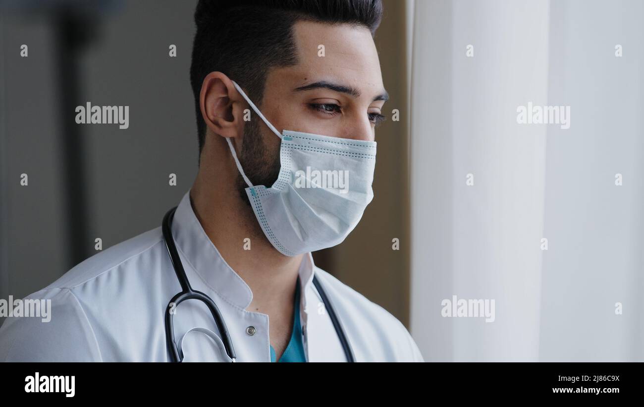 Homme pensif arabe homme thérapeute chirurgien porter blanc uniforme masque médical debout sur le lieu de travail regardant la fenêtre empêchant la propagation Banque D'Images