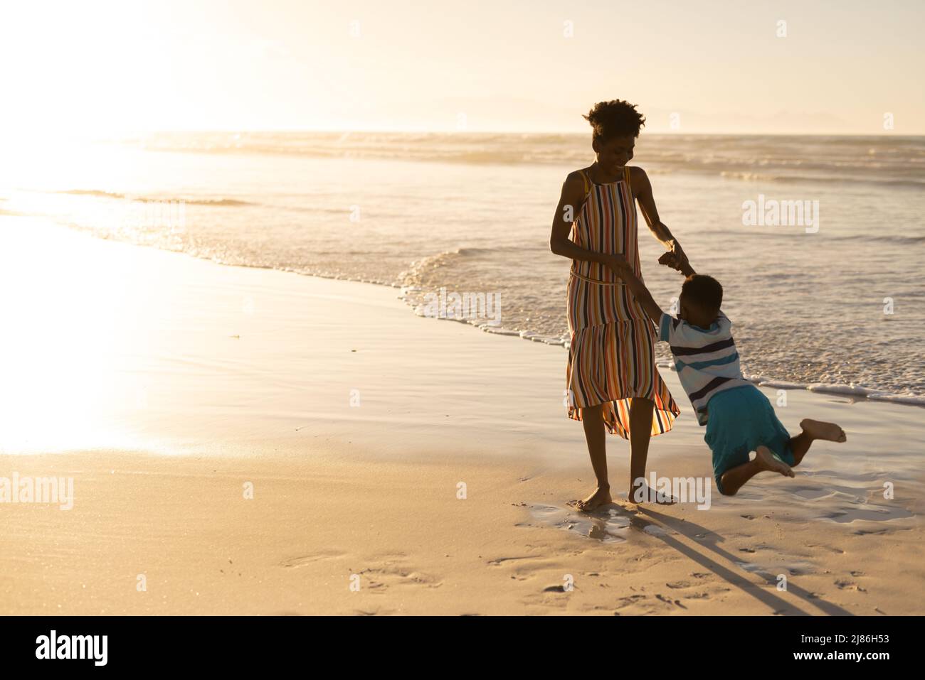 Joueuse jeune femme afro-américaine tenant les mains de son fils et lui faisant tourner à la plage contre le ciel Banque D'Images