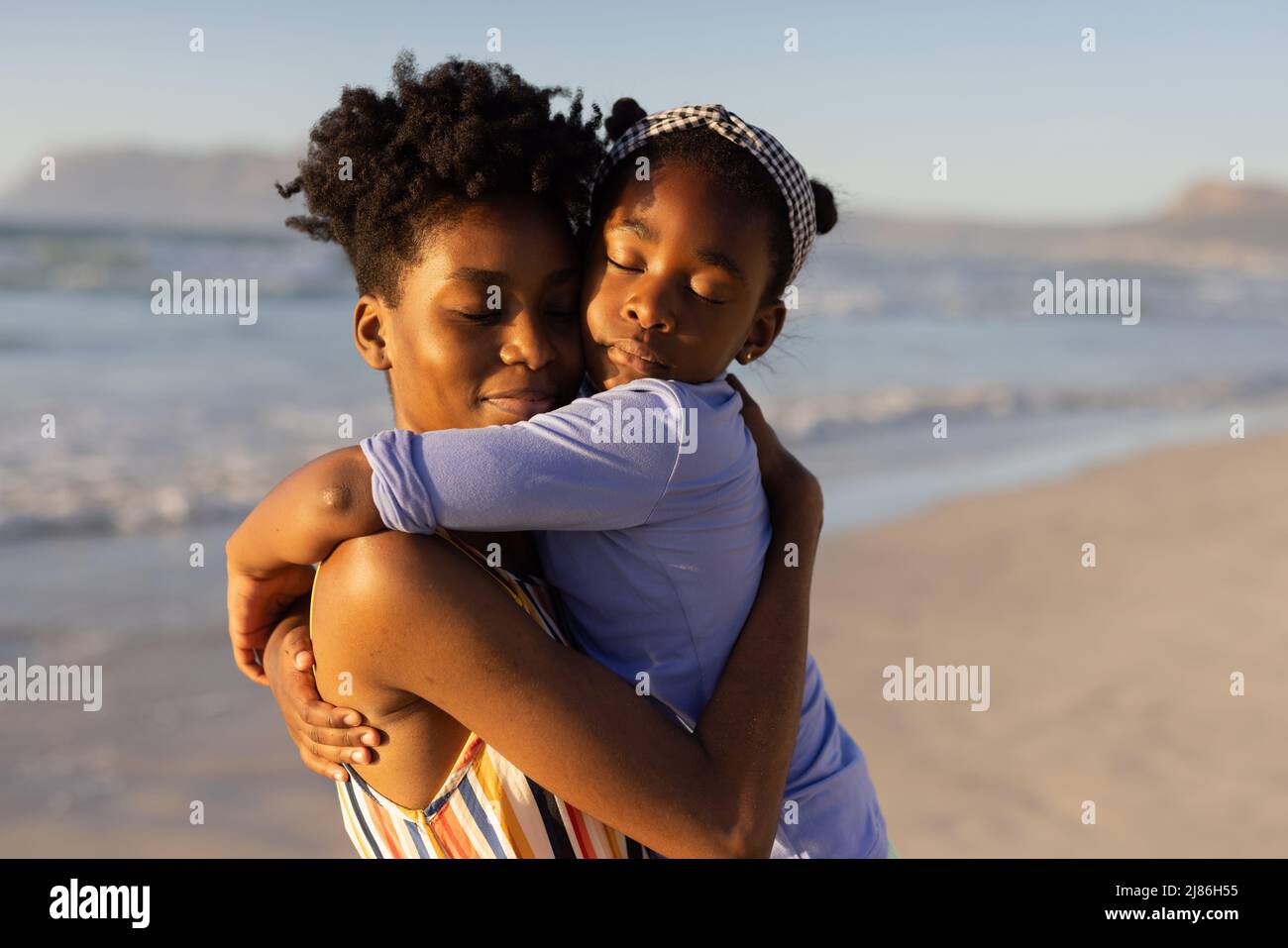 Jeune femme afro-américaine avec les yeux fermés embrassant la fille contre la mer et ciel clair au coucher du soleil Banque D'Images