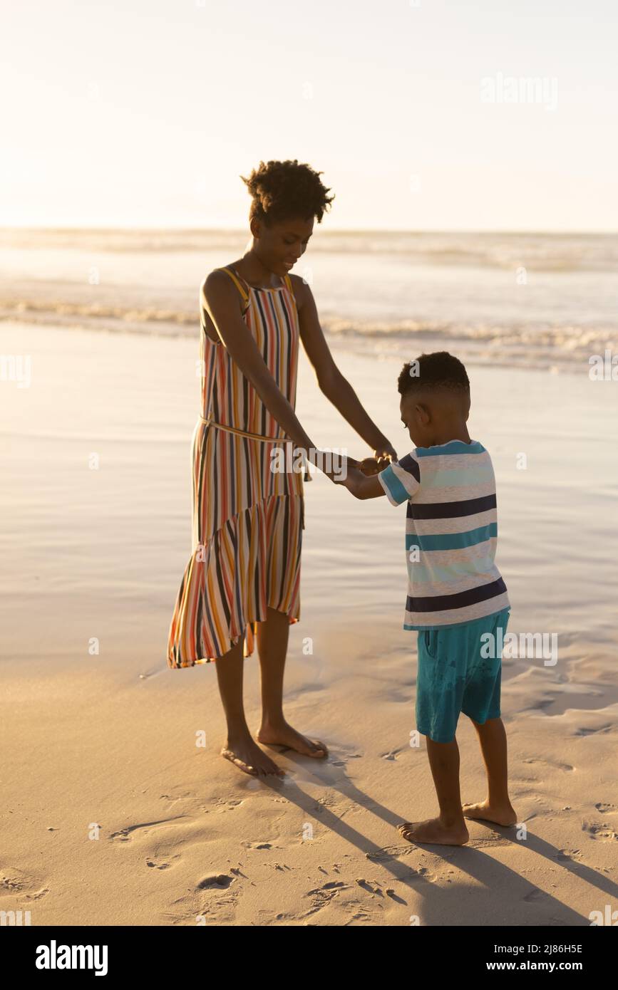 Jeune femme afro-américaine tenant les mains de son fils tout en se tenant sur la plage contre le ciel clair au coucher du soleil Banque D'Images