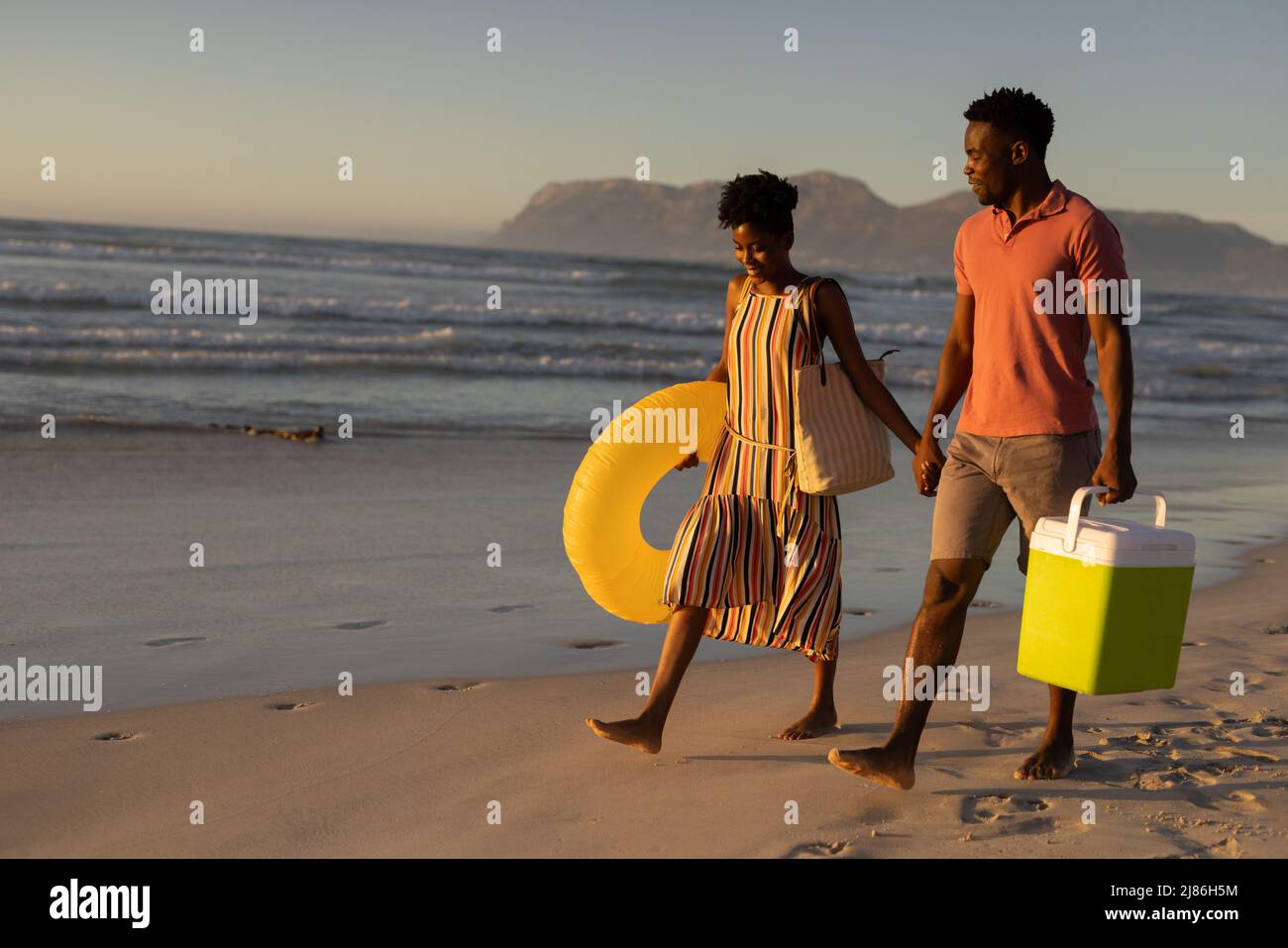 Jeune couple afro-américain avec glacière et anneau gonflable tenant les mains tout en marchant sur la plage Banque D'Images