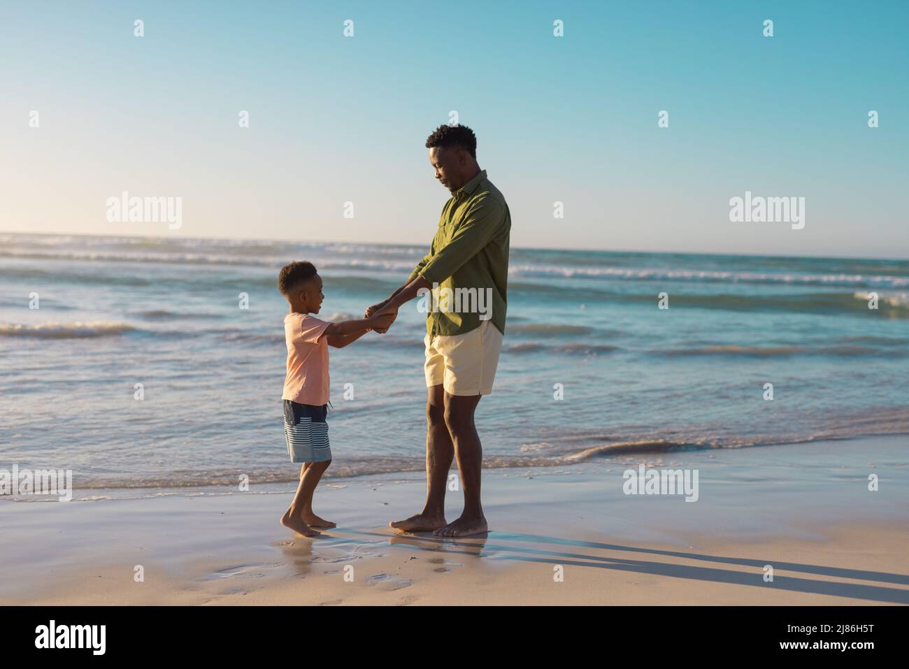 Jeune Afro-américain tenant les mains de son fils tout en se tenant à la plage contre le ciel clair au coucher du soleil Banque D'Images