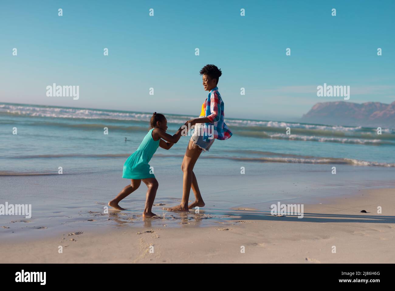 Vue latérale de la jeune femme afro-américaine tenant les mains de sa fille et jouant à la plage contre le ciel Banque D'Images