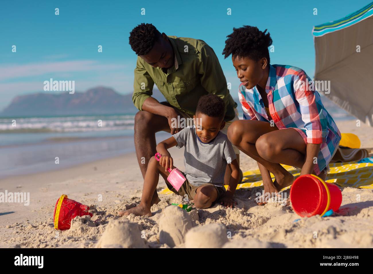 Jeunes parents afro-américains regardant son fils jouer avec du sable à la plage contre le ciel par beau temps Banque D'Images