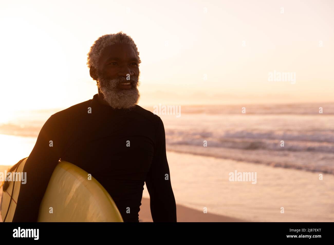 Homme senior afro-américain barbu avec planche de surf regardant loin contre la mer et ciel clair au coucher du soleil Banque D'Images