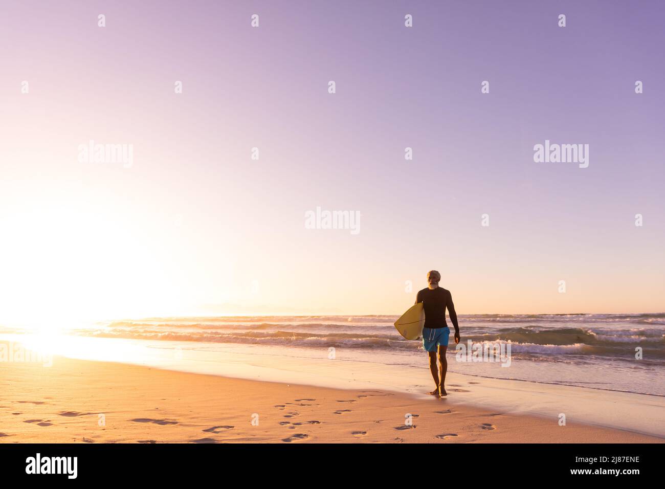 Homme senior afro-américain avec planche de surf marchant contre un paysage marin et ciel clair au coucher du soleil Banque D'Images