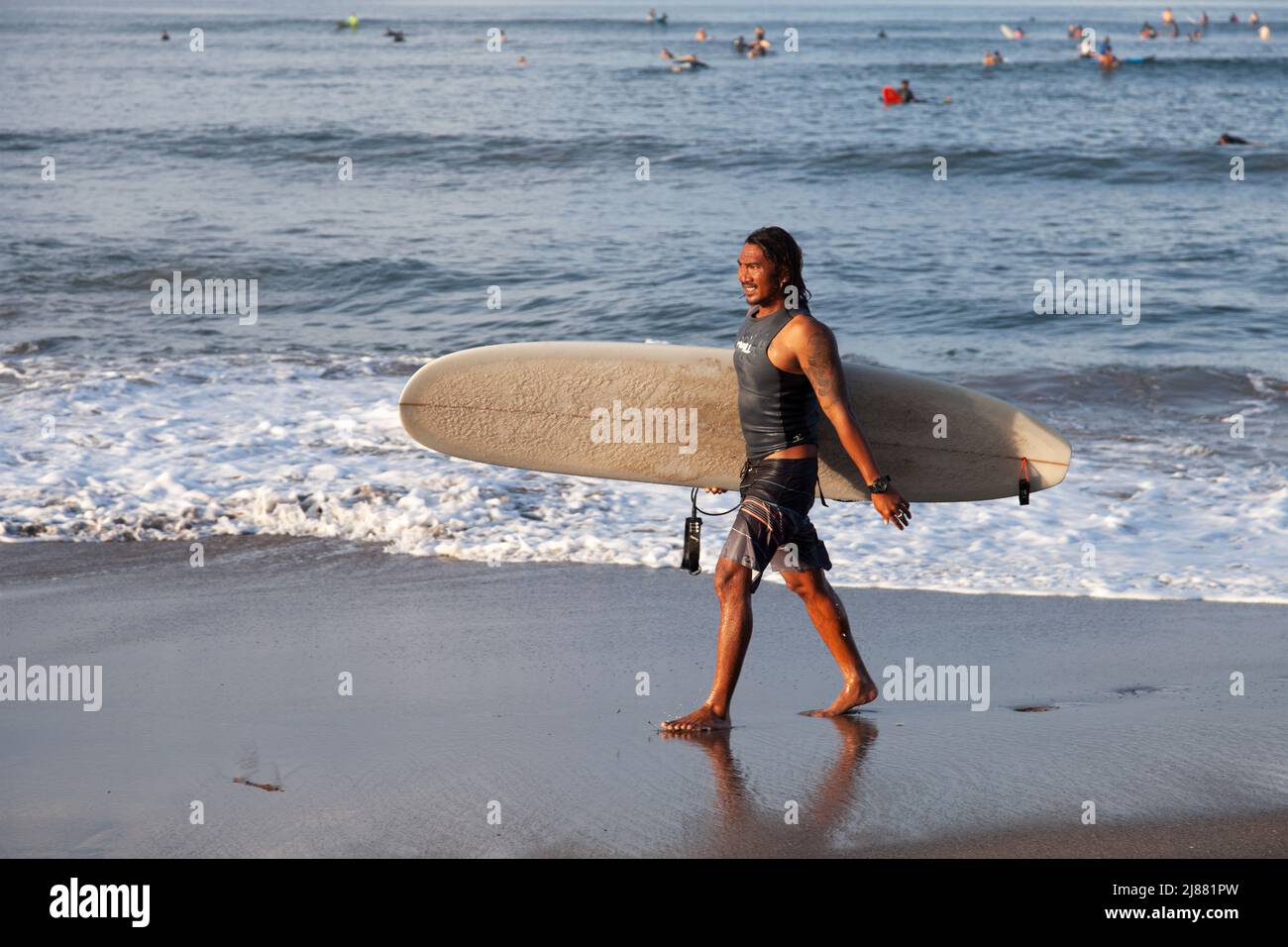 Un surfeur indonésien local marchant le long de la plage de Batu Bolong à Canggu, Bali, Indonésie portant une planche de surf blanche avec des surfeurs dans la mer derrière, Banque D'Images
