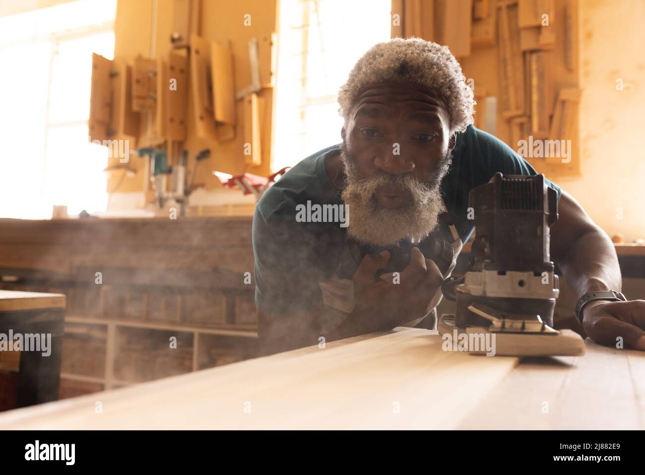 Charpentier africain américain mature soufflant de la sciure tout en utilisant la ponceuse à l'établi dans l'atelier Banque D'Images