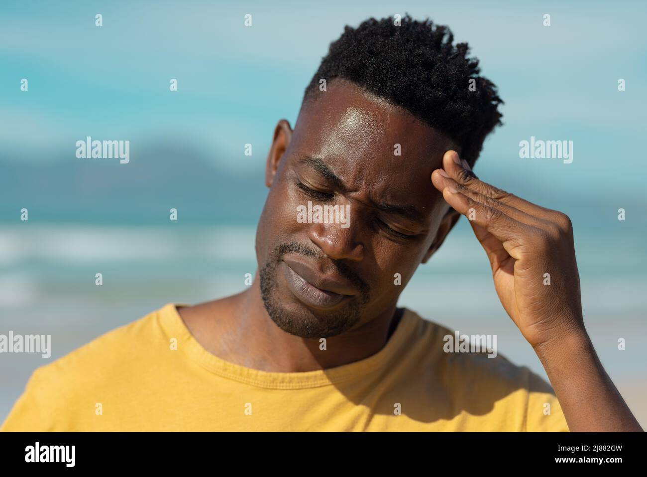 Gros plan du jeune homme afro-américain avec les yeux fermés, touchant la tête à la plage contre le ciel en été Banque D'Images