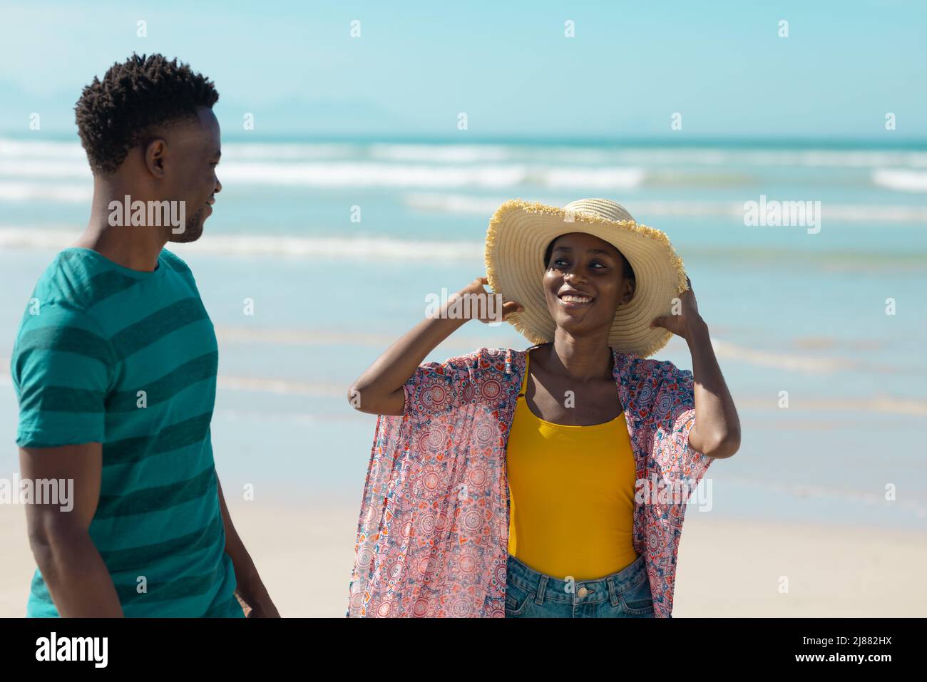 Jeune femme afro-américaine souriante portant un chapeau et une veste sarong regardant un petit ami à la plage Banque D'Images
