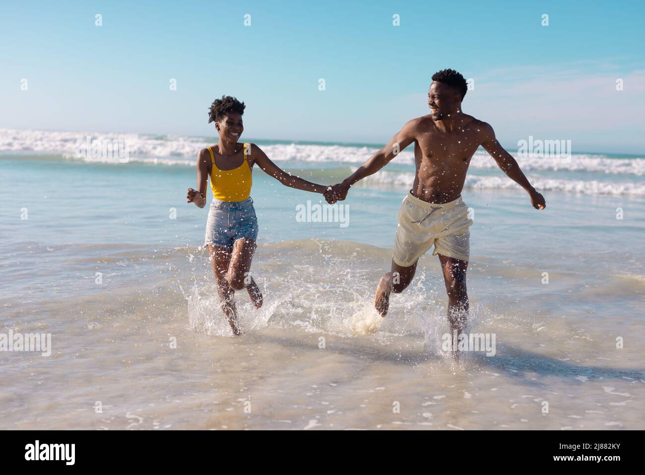 Un jeune couple afro-américain insouciant se tient les mains et court dans la mer contre le ciel bleu Banque D'Images