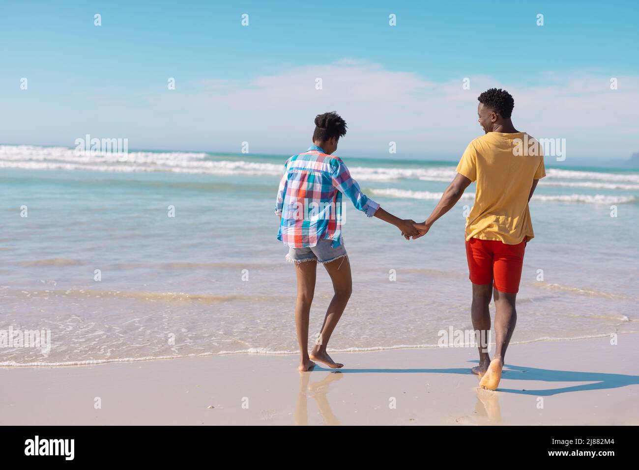 Vue arrière d'un jeune couple afro-américain tenant les mains et marchant sur la mer contre le ciel Banque D'Images