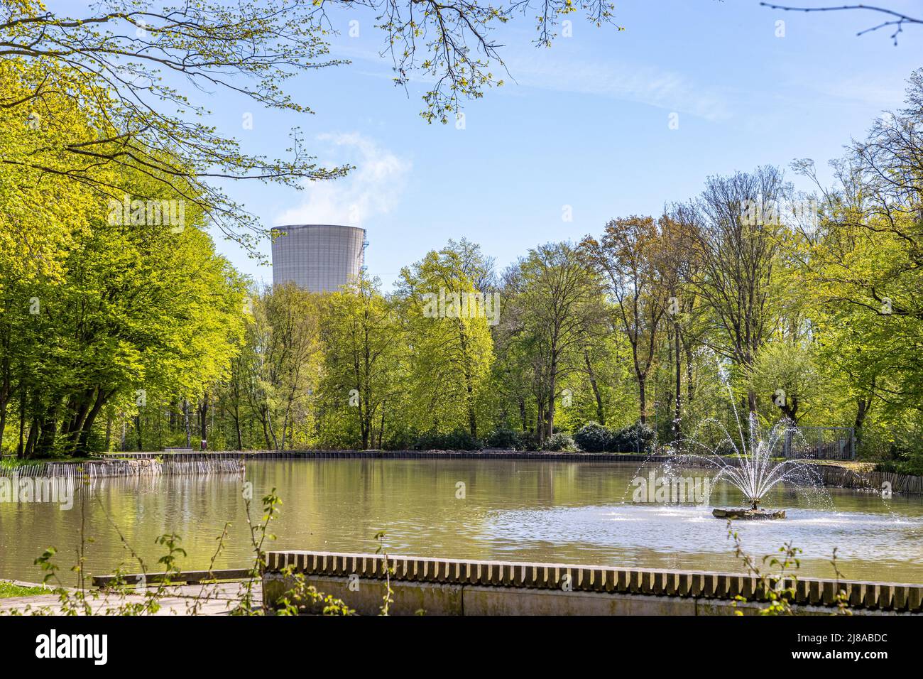 Étang avec une petite fontaine, arbres verts luxuriants et une énorme cheminée d'un complexe industriel en arrière-plan, jour ensoleillé avec un ciel bleu de source à Heidek Banque D'Images