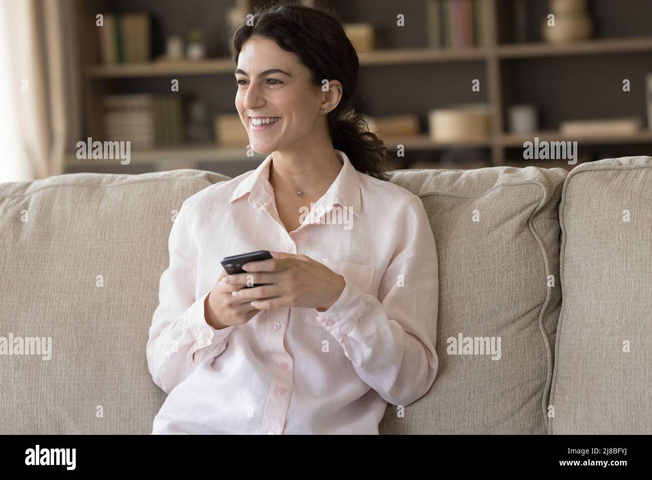 Une jeune femme assise sur un canapé tient le sourire d'un smartphone sur le côté Banque D'Images