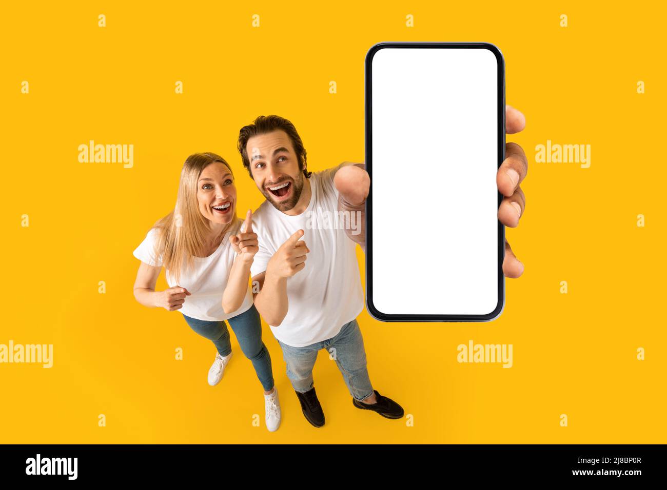 Heureux surpris jeune couple européen excité en t-shirts blancs montrent les pouces vers le haut au smartphone avec écran blanc Banque D'Images