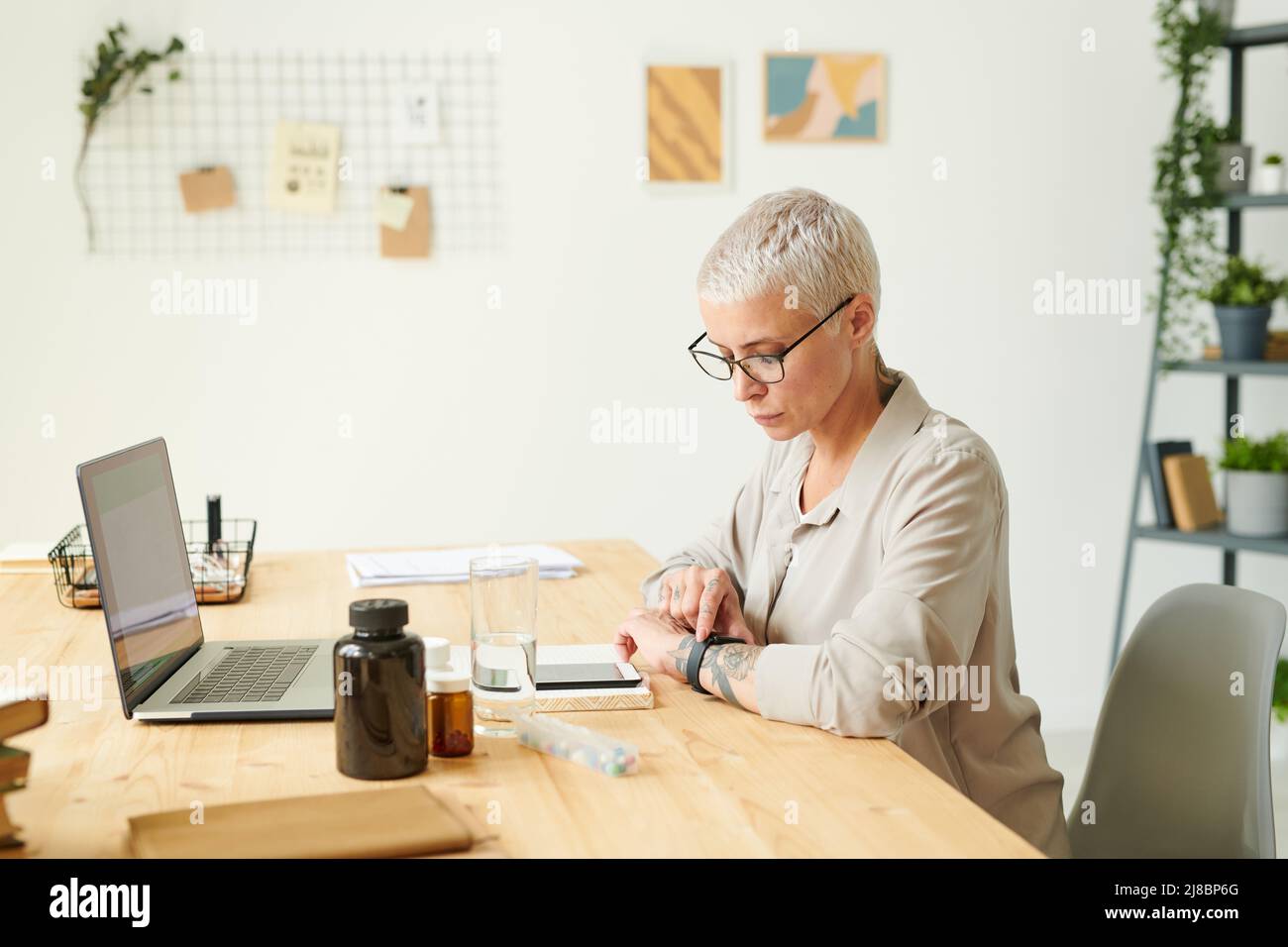 Femme moderne sérieuse avec des cheveux blonds vérifier le temps avant de prendre des vitamines au bureau, concept de biopiratage Banque D'Images