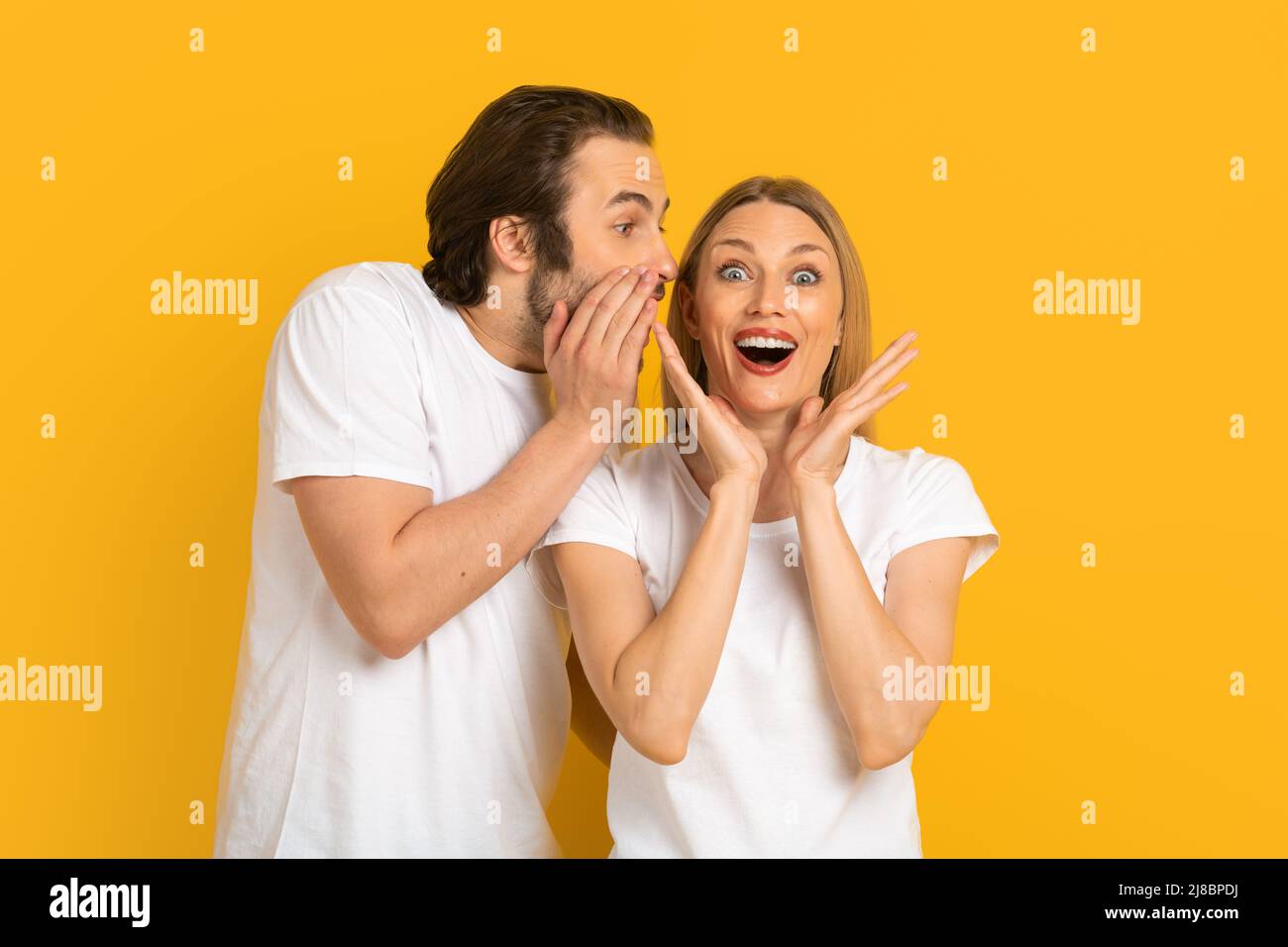 Le jeune mari européen en t-shirt blanc murmure secret à l'oreille de sa femme surprise à bouche ouverte Banque D'Images