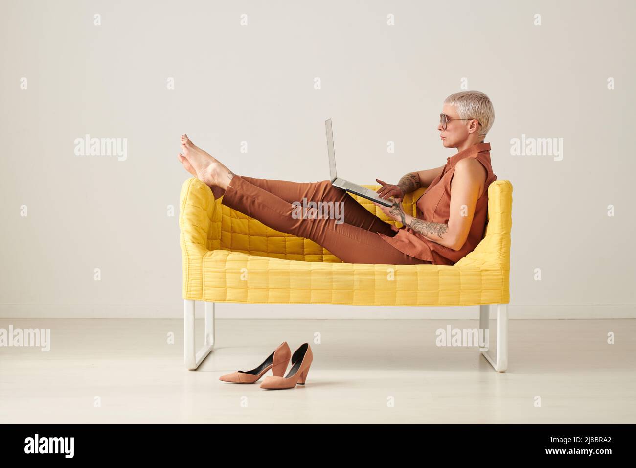 Femme d'affaires sérieuse détendue avec cheveux blond court assis sur un canapé jaune et les sites de surf sur ordinateur portable Banque D'Images