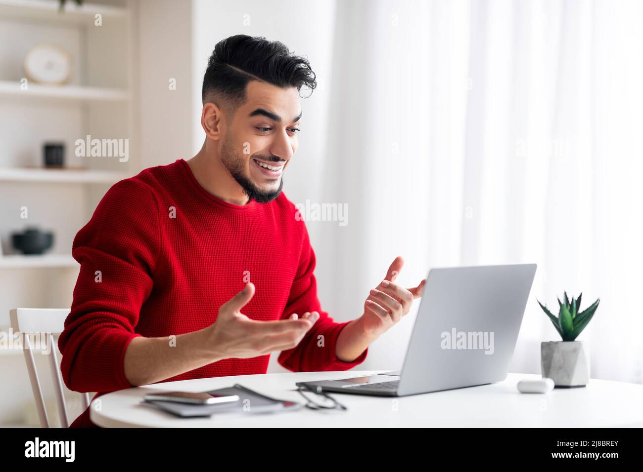 Souriant surpris jeune homme moyen-oriental avec la barbe regarde l'ordinateur et gestuelle au travail à la maison Banque D'Images