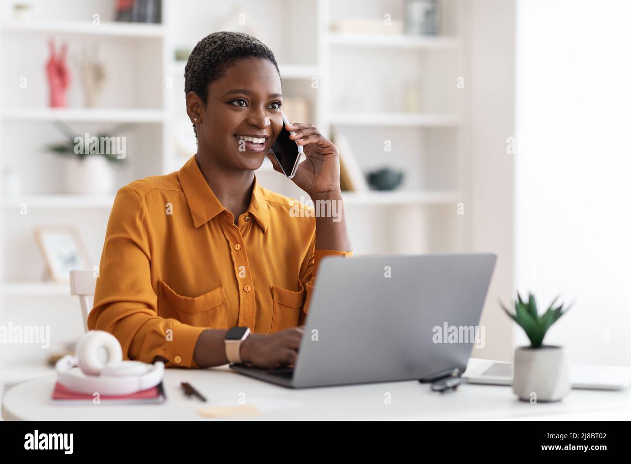 Femme noire assise à une table avec un ordinateur portable, ayant une conversation téléphonique Banque D'Images
