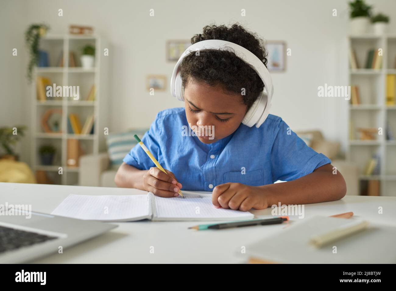 Un lycéen afro-américain sérieux dans des écouteurs assis à son bureau et prenant des notes tout en écoutant un tuteur en ligne Banque D'Images